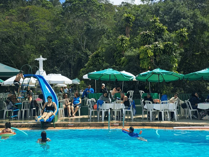 Piscina ao ar livre com tobogã azul, guarda-sóis verdes e a mata ao fundo